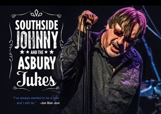 Southside Johnny stands on stage holding his microphone while leaning into the mic stand. His Western style logo is to his left.